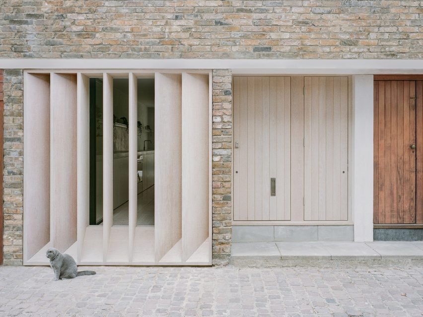 Exterior de una casa de ladrillo con una puerta de madera, persianas de madera frente a una ventana grande y un gato gris