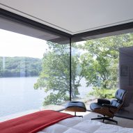 Bed, lounge chair and foot stool in a bedroom with a large corner window overlooking a lake