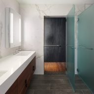 A bathroom with dark tiled flooring, white marble walls and white basins with wood drawers