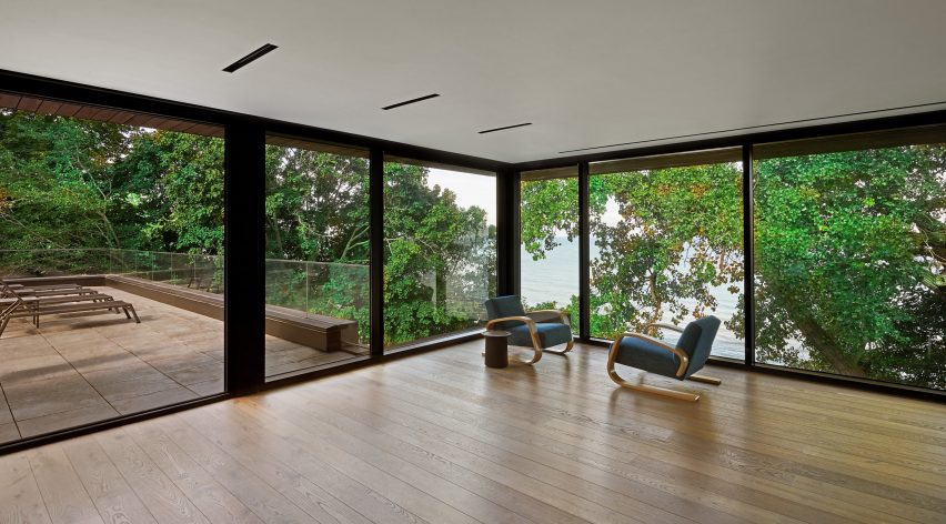 A room with wooden flooring, floor-to-ceiling windows and two lounge chairs overlooking trees