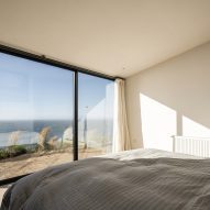 Bedroom with white walls, a bed with grey bedding and floor-to-ceiling windows overlooking the sea