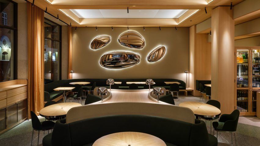 A restaurant interior with wood columns and beams, wood tables, green up،lstered seating and oval metallic wall lights