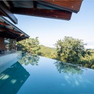 A home with glass walls next to an infinity pool overlooking treetops