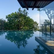 A home with glass walls next to infinity swimming pool overlooking treetops