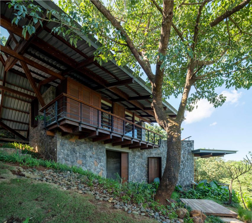 Exterior of Lateral Verandah House in India on a grassy slope with stone and timber walls