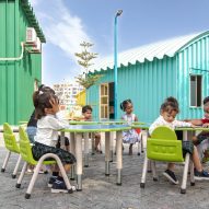 Children sitting at outdoor tables and chairs with blue and green metals buildings behind