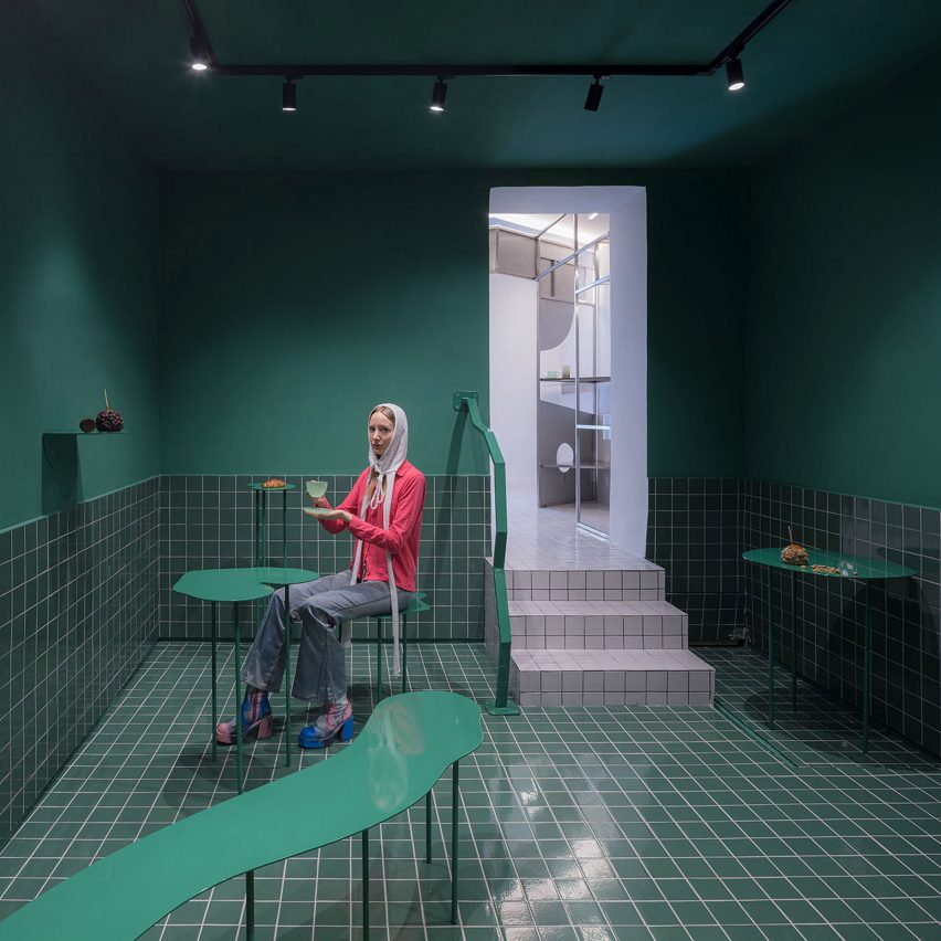 A dark green room with green floor tiles, curved green metal tables and a person sitting with a coffee and croissant