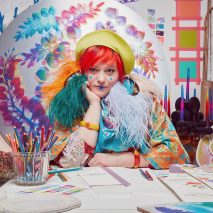 Person with red hair and green beret sitting at a table with colouring pencils