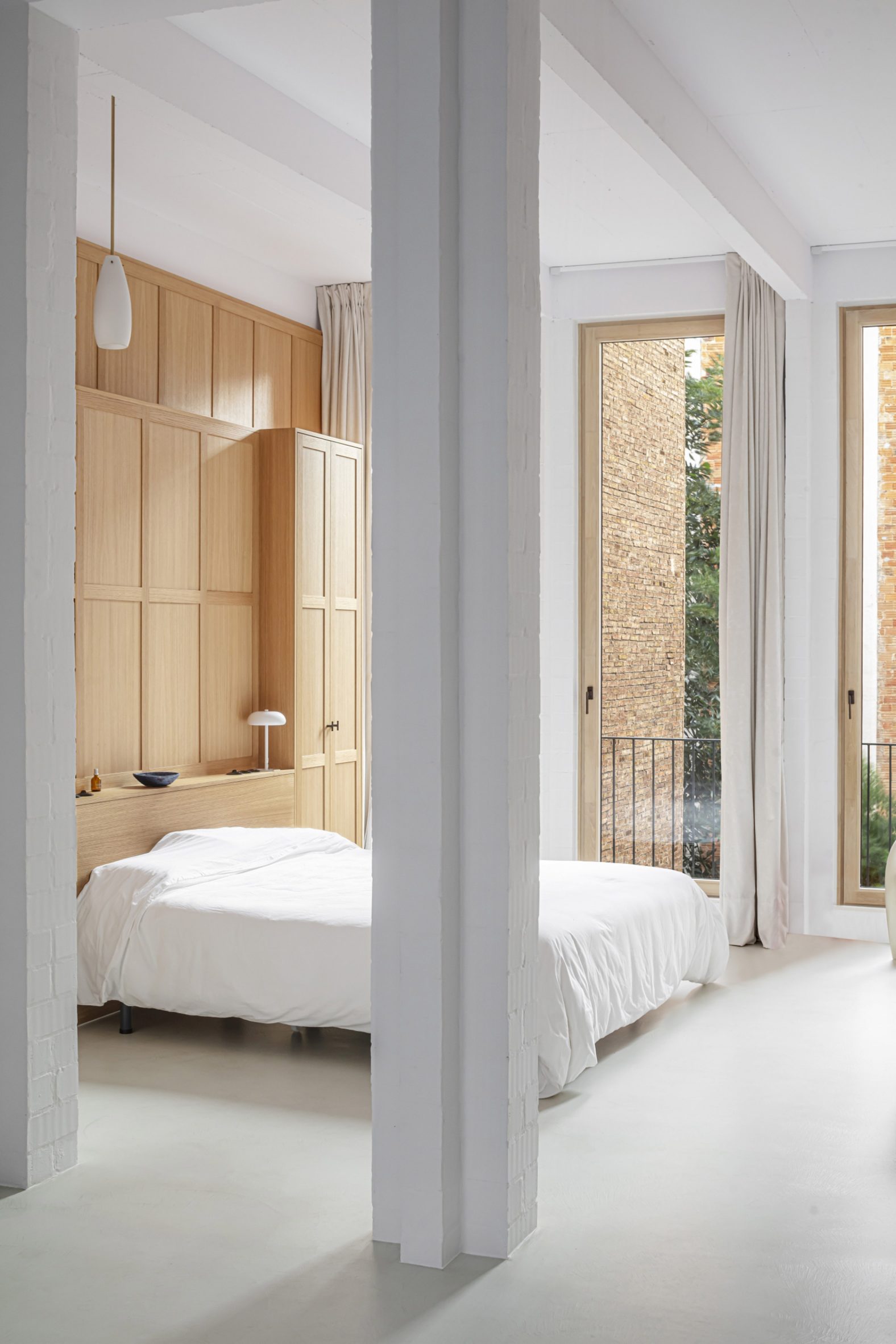 Interior of a bedroom with white floors, white square columns, wood-panelled wall and a bed with white bedding