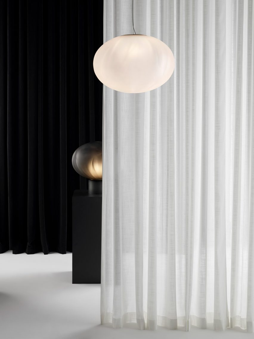 Photo of a frosted glass Méne pendant hanging in front of a partially open white curtain, with a Méne table lamp in frosted black on a black plinth behind it