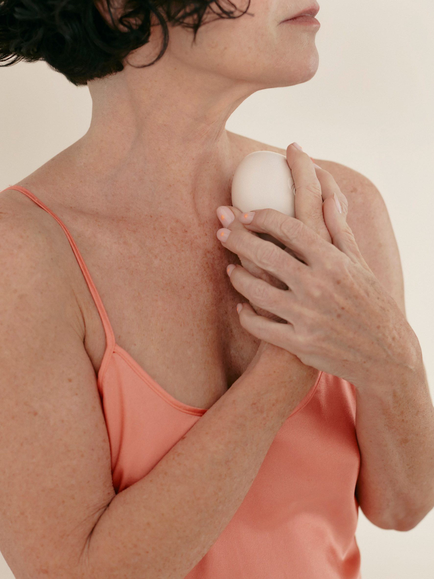 Photo of a middle-aged woman in a coral pink camisole, holding the Tides massager to her chest
