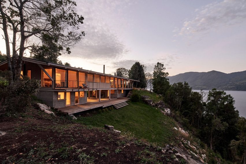 Timber house on a hillside with large windows revealing warmly-lit rooms