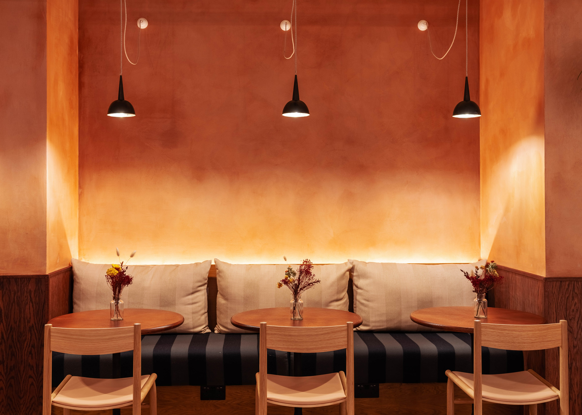 Restaurant interior with an orange ombre wall, black pendant lights, bench seating and wooden tables and chairs