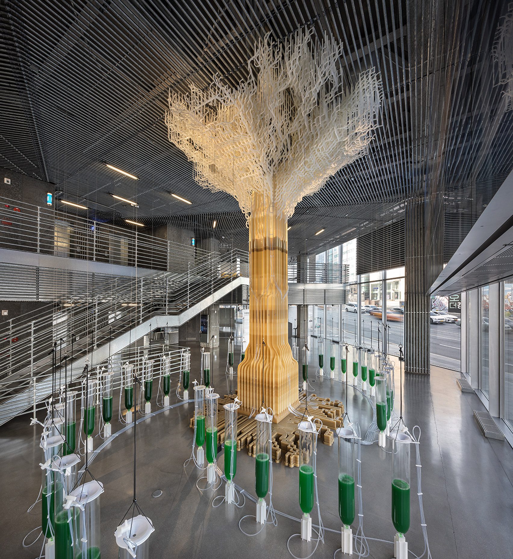 Photo of a 10-metre-tall yellow-brown tree sculpture sitting within the foyer of a modern corporate building, surrounded by clear glass vessels full of green liquid