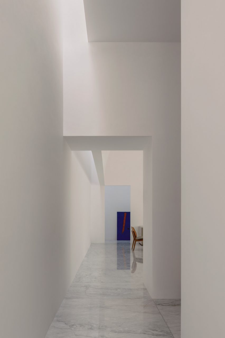 Koridor interior putih dengan bukaan persegi panjang mengarah ke ruang tamu dengan lukisan biru