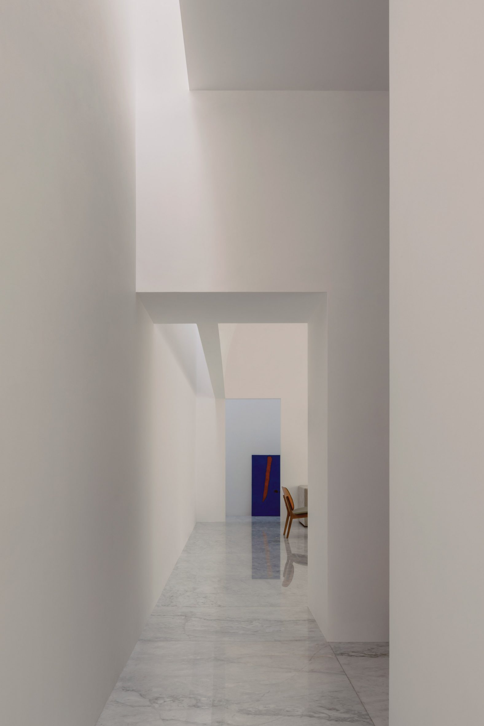 A white interior corridor with a rectangular opening leading to a living room with a blue painting