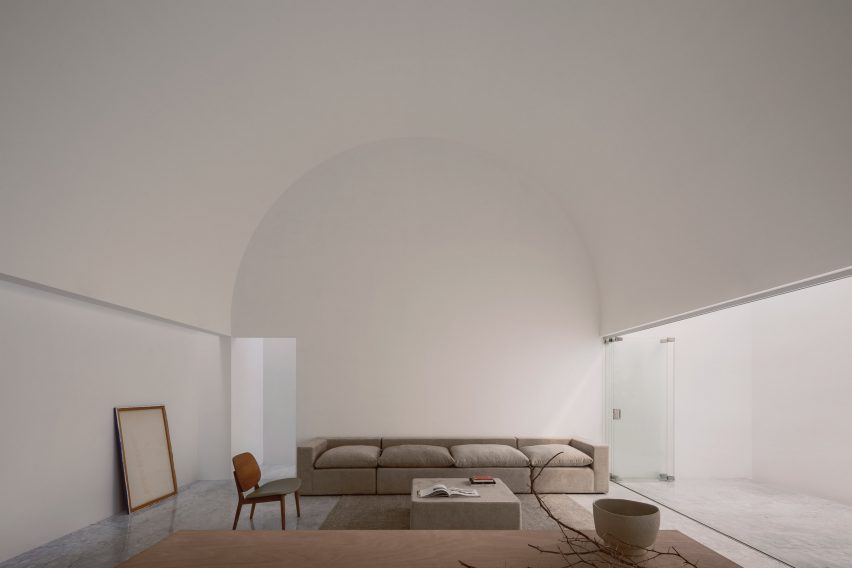 A white room with a vaulted ceiling, a gray sofa, a gray rug, a coffee table and glass doors to one side leading to the outdoors