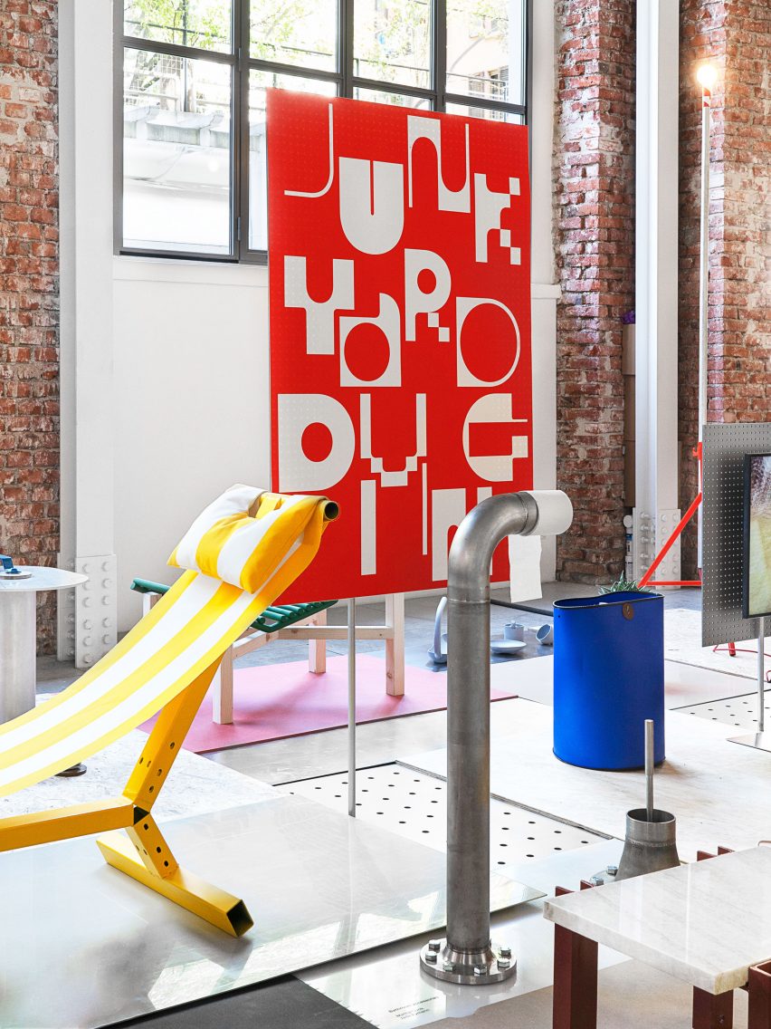 A yellow hammock-style chair with a metal base, a toilet roll holder made from a metal pipe and a blue bag sit on display at an exhibition with a red sign reading Junkyard Diving behind them