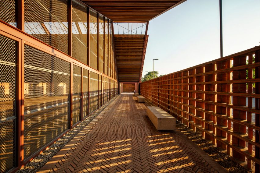 Una pasarela exterior cubierta con suelo de ladrillo y paredes de madera junto a un edificio acristalado con dos aguas