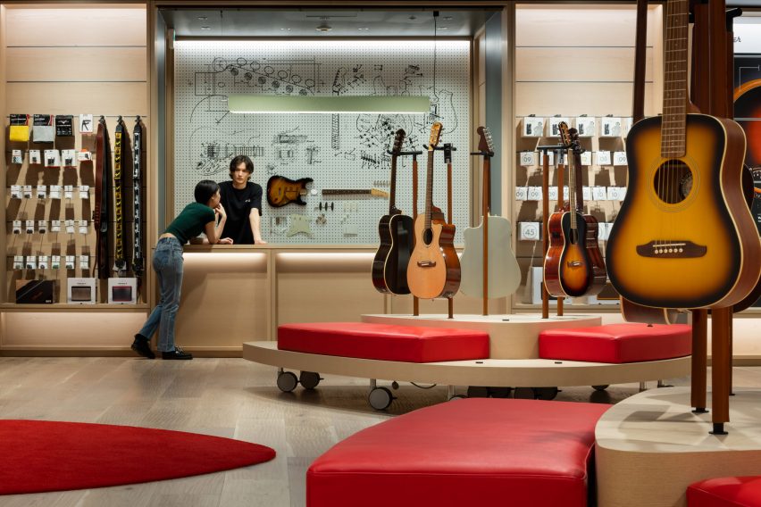 Photo of a room with light-coloured walls, red cushioned bench seating and displays of acoustic guitars in the centre of the room, and racks full of guitar accessories at the back. Near the back wall, a man stands behind a counter with a display of guitar parts behind him and talks to a woman who is leaning on the front of the counter