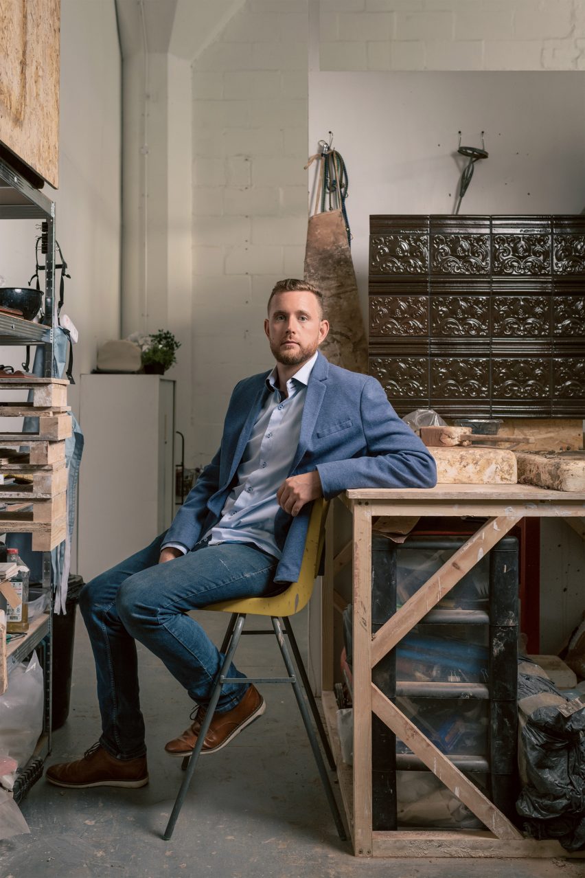 Photo of designer Jeffrey Miller sitting in his studio in front of a small wall of brown tiles he has made from London Underground waste