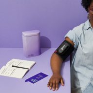 Photo of a Black woman with a blood pressure cuff on her right arm, which rests on a table. Spora Health's Momma's Kit tin also sits on the table, along with an open notebook with prompts printed on its pages