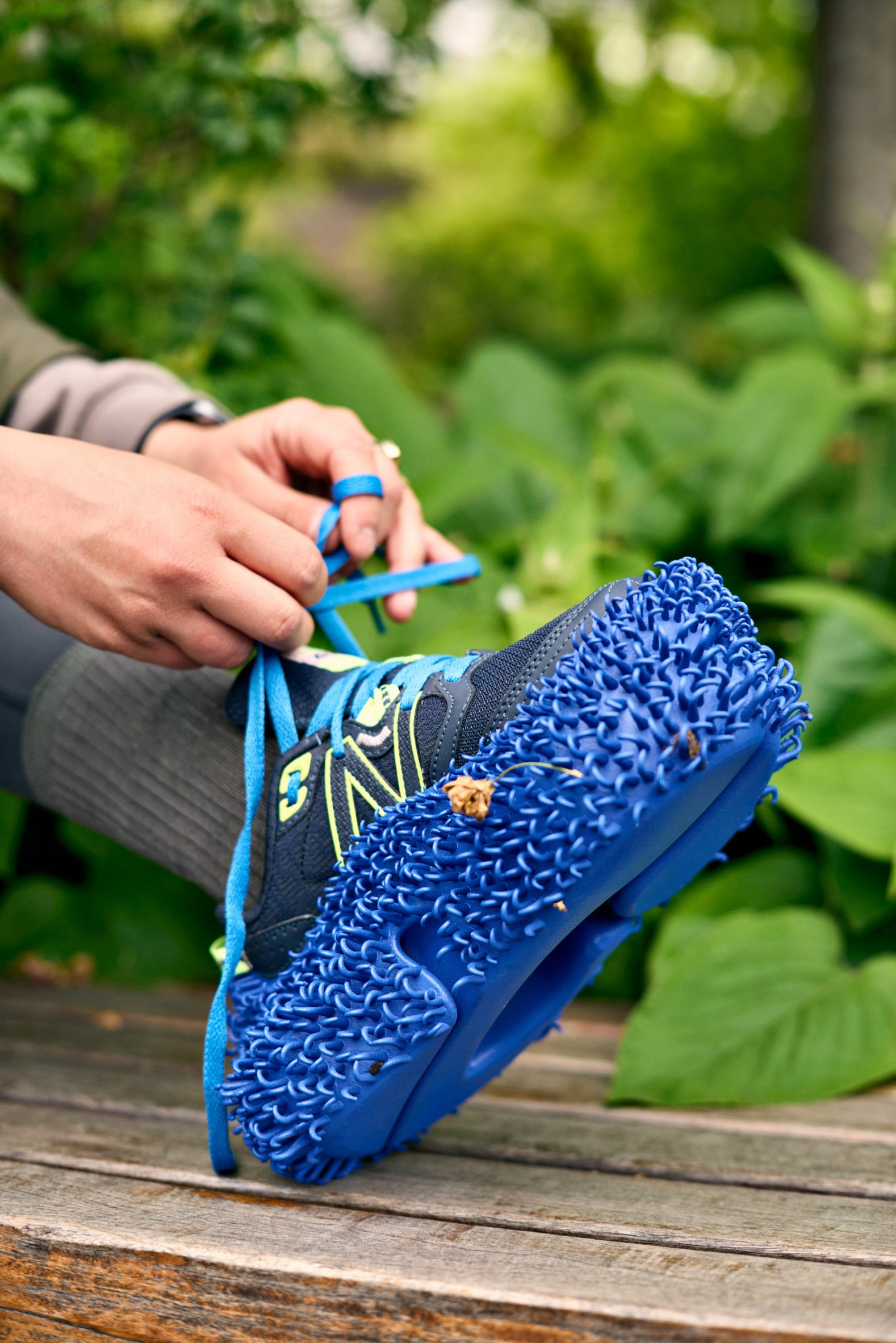Photo of a running shoe with a chunky, bright blue plastic sole covered in tiny hooks like hair, while a pair of hands ties the laces