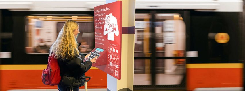 Girl interacting with a red heart health analysing poster in a train station