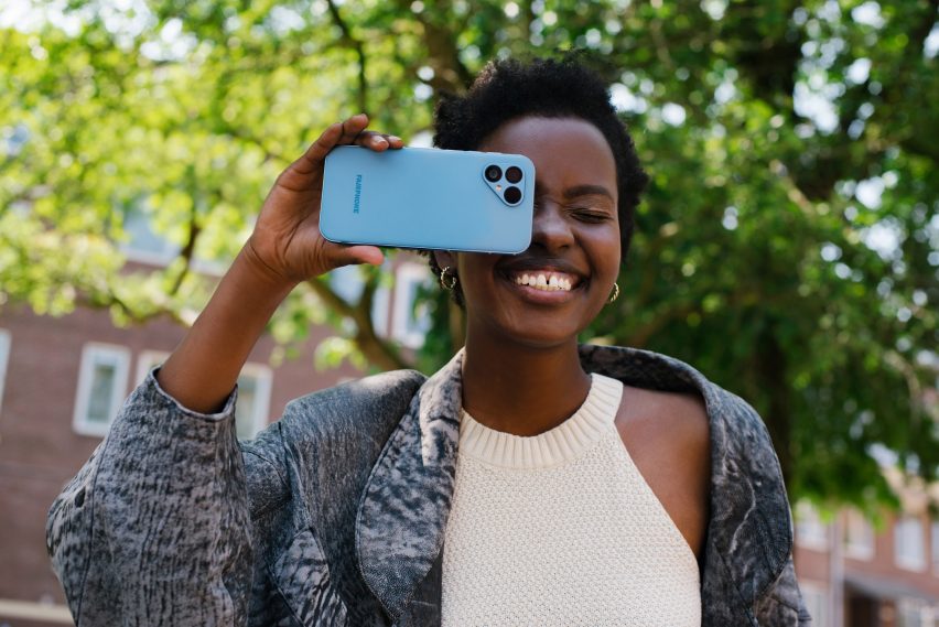 Retrato de una joven sonriente sosteniendo un Fairphone 5 azul en el ojo 