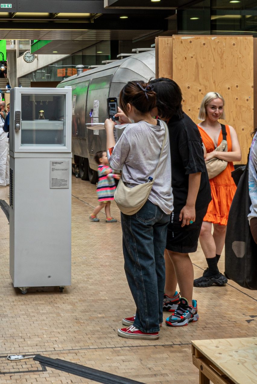 Photograph of visitors to the Central Saint Martins graduate exhibition taking a photo on a phone of a tall white fridge displaying a cup of ice-cream