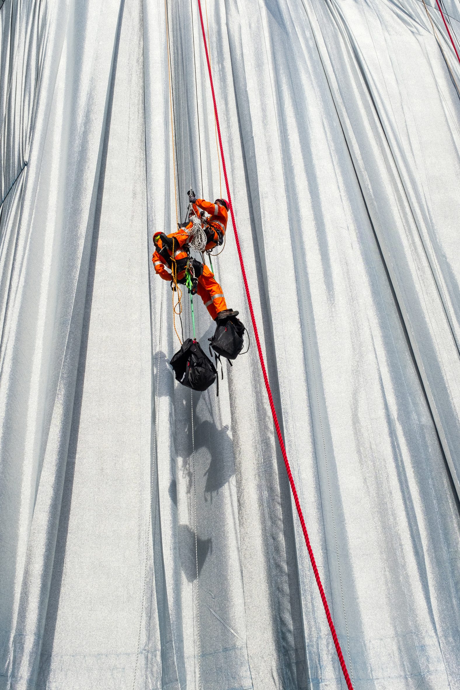 Photo of a two workers in high-vis gear hanging on the outside of the Christo and Jeanne-Claude's L'Arc de Triomphe Wrapped installation, showing the red ropes and silvery fabric up close