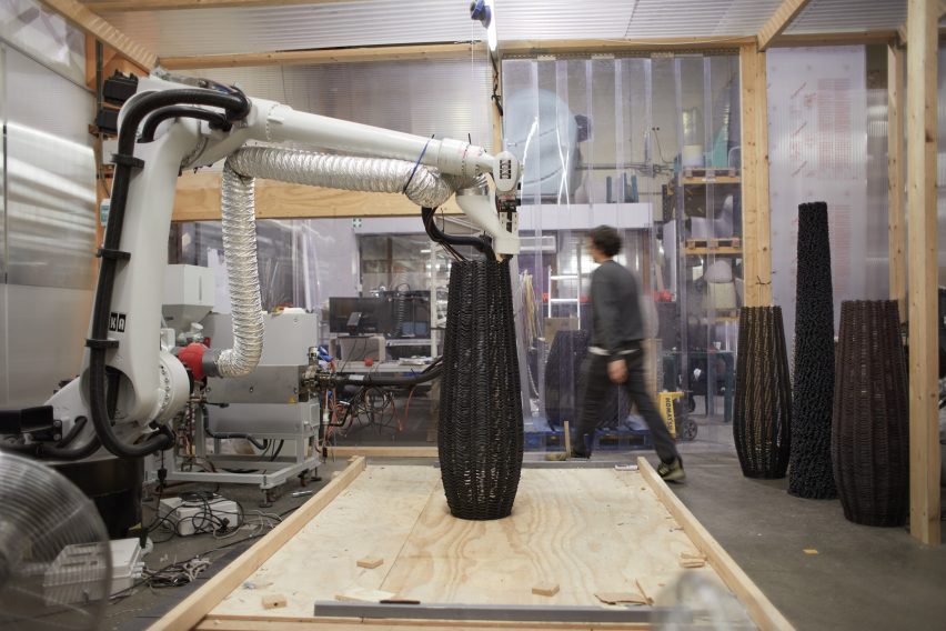 Фотография промышленного робота в мастерской, изготавливающего сосудоподобную форму из черного полимера, который он экструдирует.
