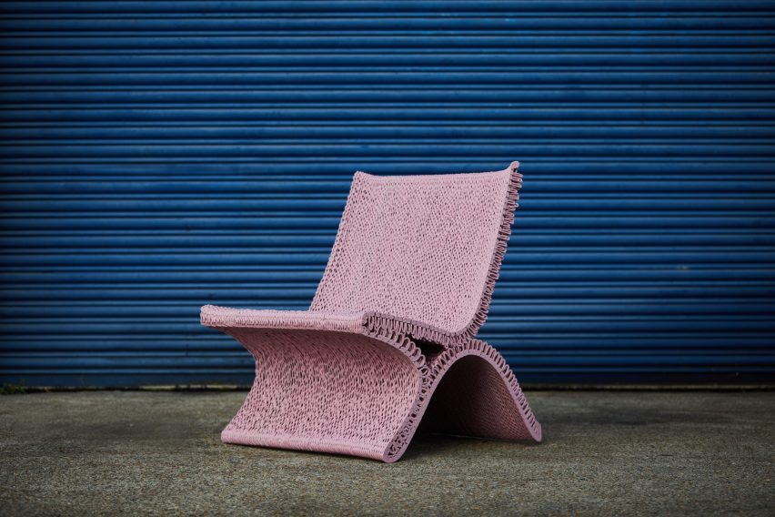 Фотография розового стула, распечатанного на 3D-принтере в петлях из пластикового шнура, на бетонном тротуаре перед синей роллетной дверью.
