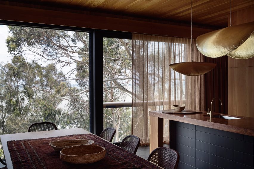 Деревянная кухня с деревянным столом и кухонным островом, а также большими окнами с видом на верхушки деревьев.