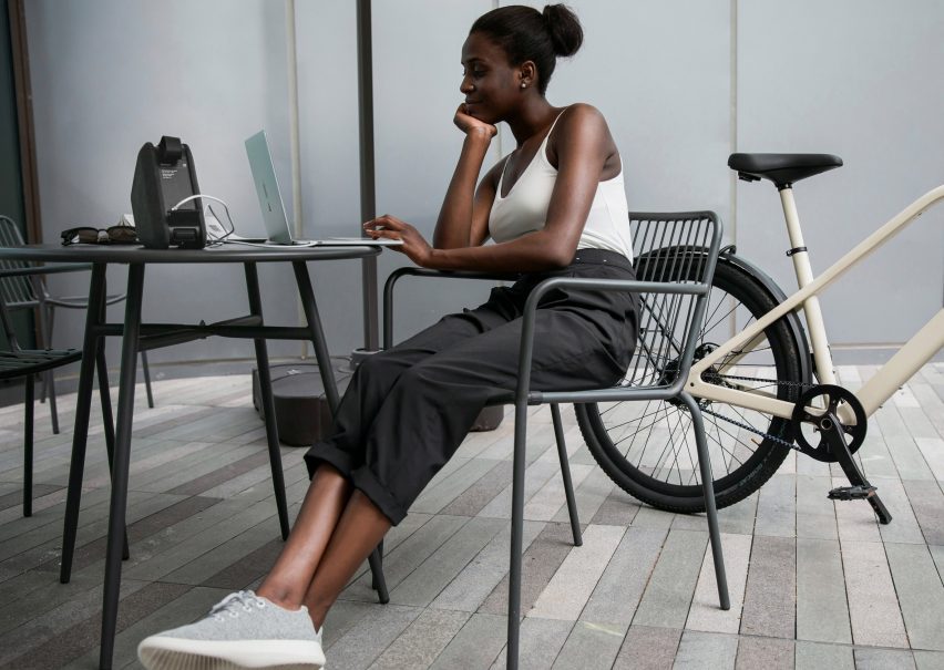 Photo of a woman at an outdoor table with her bike partially out of frame beside her and a battery pack powering her laptop on the table