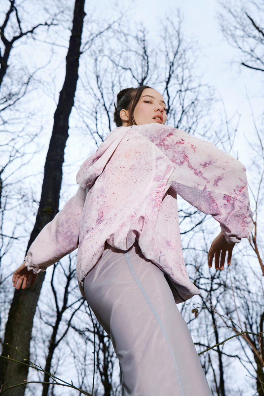 Campaign photo of a female model wearing the Exploring jacket in a forest. The jacket has been dyed by bacteria to have a patchy pink and purple pattern on white silk