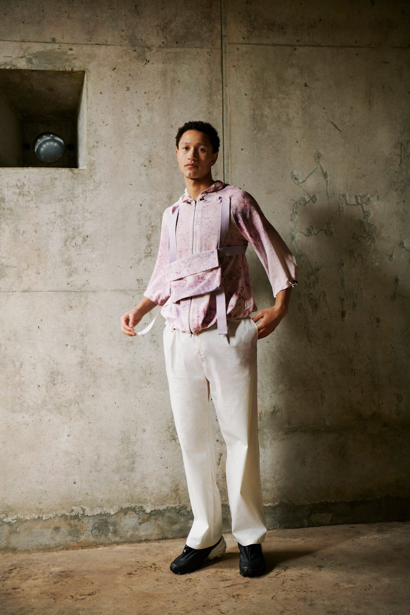 Photo of a male-presenting model wearing the pink-hued NPOL Exploring jacket and musette within an industrial environment