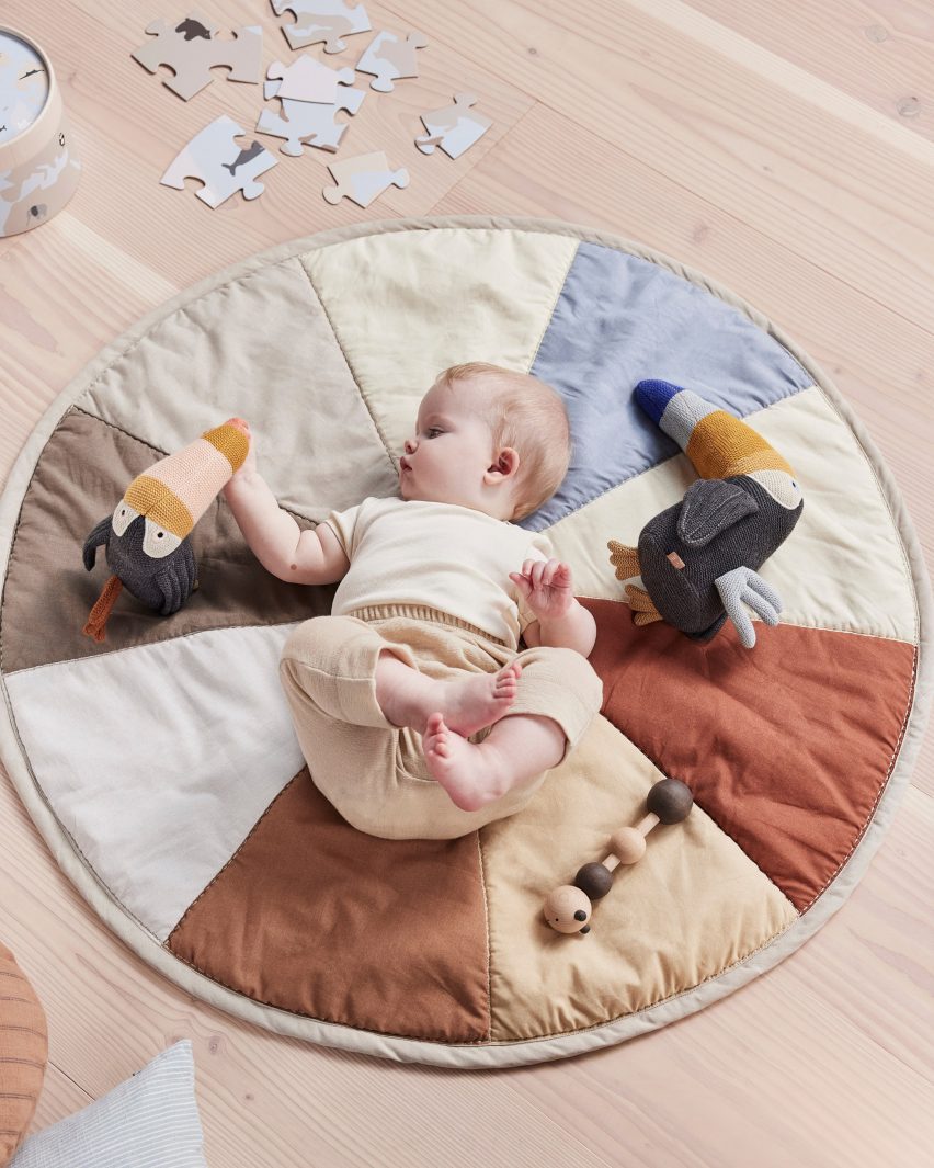 Фотография ребенка на игровом коврике OYOY Living Design с рисунком цветового круга приглушенных землистых и пастельных тонов, играющего с вязаными мягкими игрушками.
