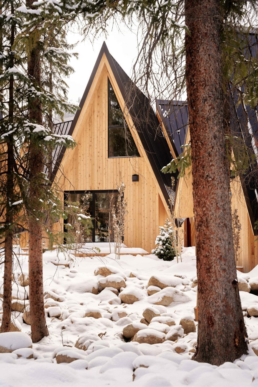 A-frame cabin seen through pine trees with snow on boulders in foreground