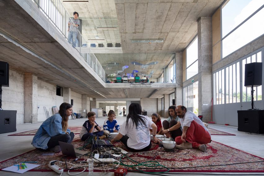 Photo of the basement level of the Wonder Cabinet in Bethlehem, showing adults and children sitting on a rug and playing with electronics while an opening in the slab above affords views into the rest of the building