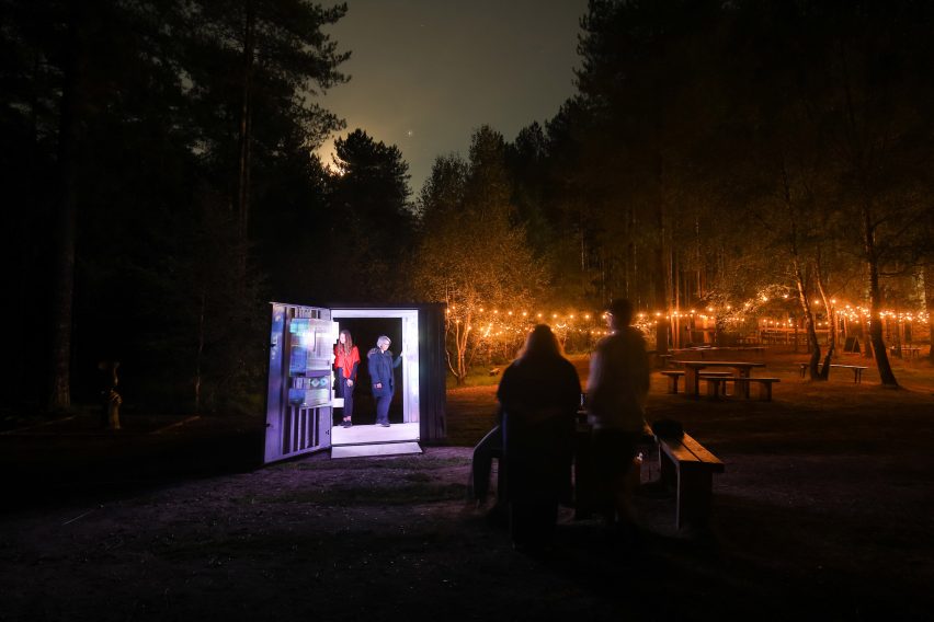 Photograph showing lit shipping container in woodland area at night