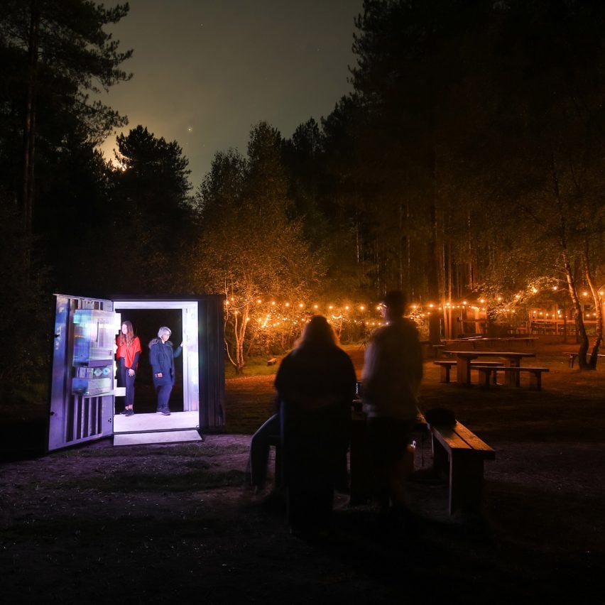 Photograph showing lit shipping container in woodland area at night