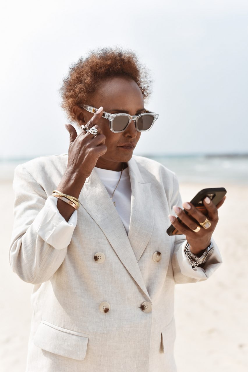 A middle-aged woman dressed in a light linen suit and sunglasses stands on a beach, looking at her phone. The finger of one hand is raised to the arm of her glasses frame