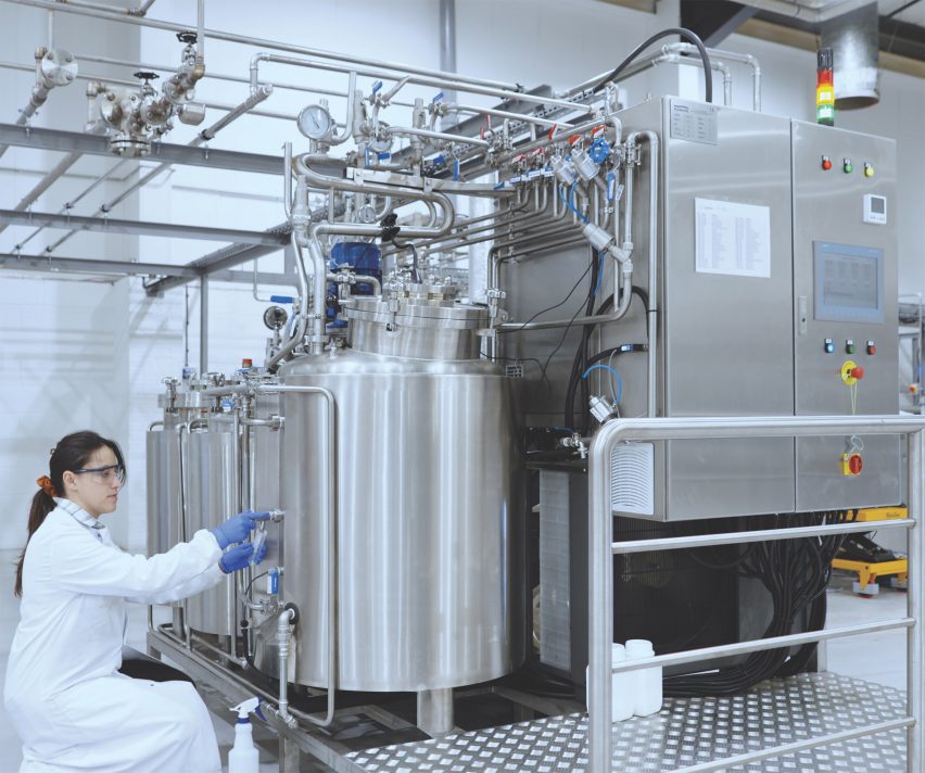 Photo of a scientist in a white lab coat tending to a row of large metal bioreactor tanks in an industrial facility