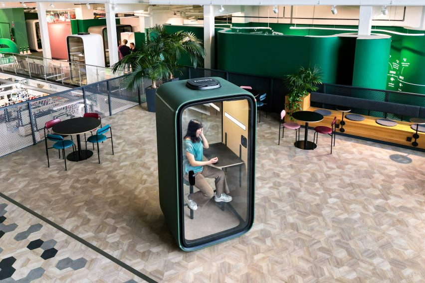 Photo or rendering of a forest green Framery One office pod within a contemporary office, placed within an otherwise open breakout space with cafe-style tables and chairs