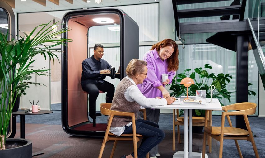 Photo of a man using the Framery One pod in an office while two women collaborate on a table outside