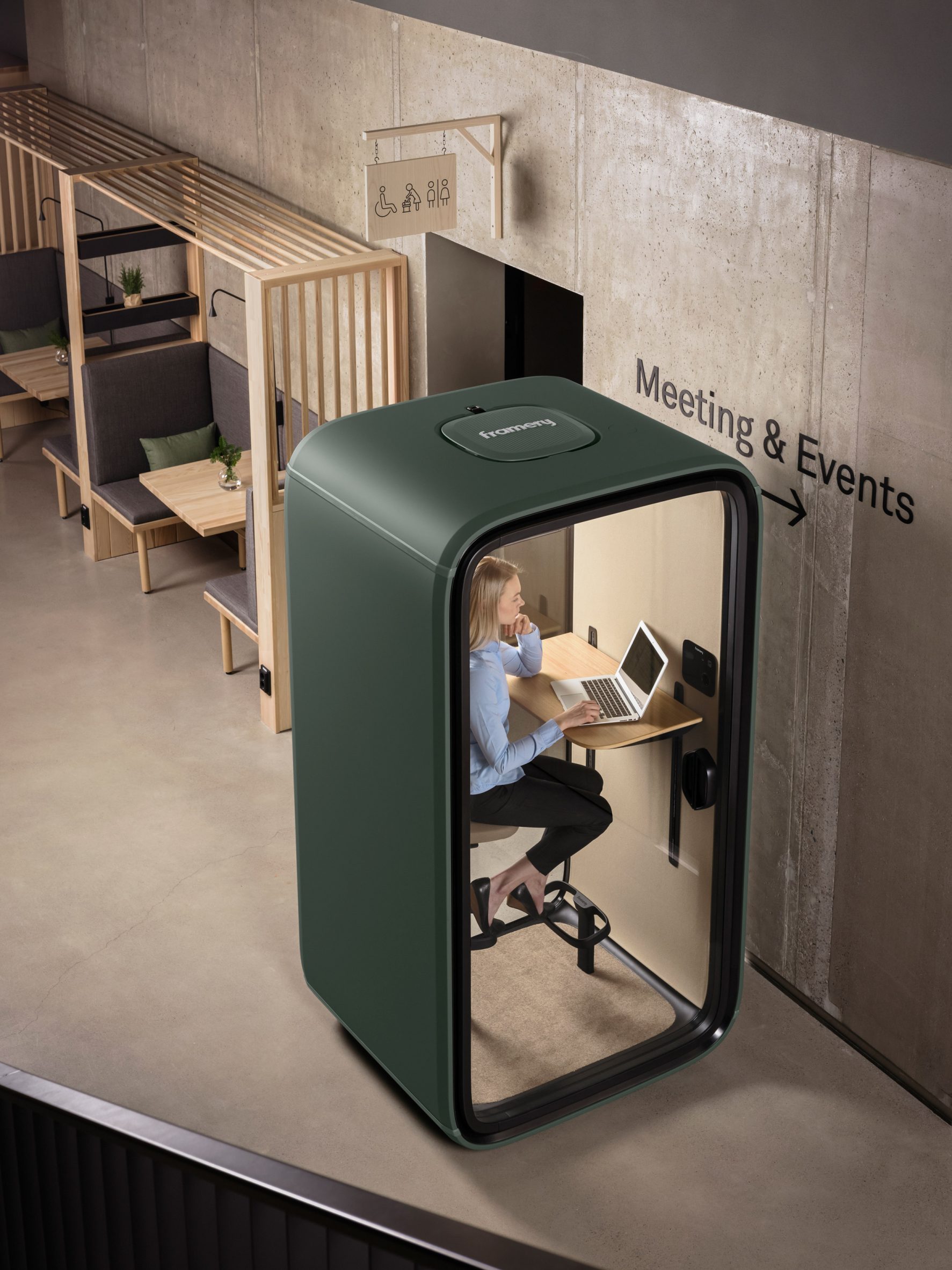 Photo of a woman sitting within a forest green-coloured single-person office pod working at a laptop. The pod sits within a modern office space surrounded by open workstations