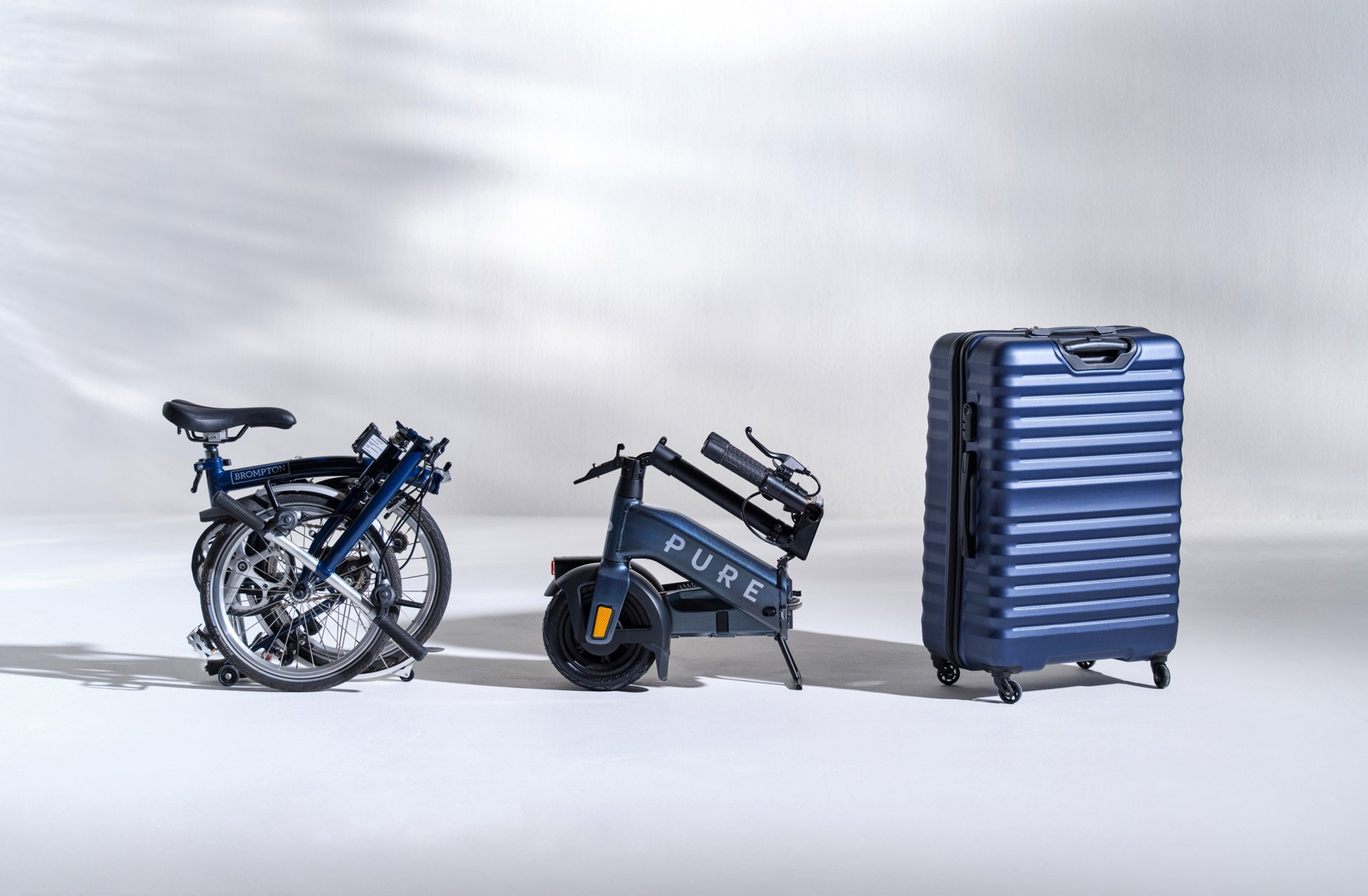 Size comparison of foldable electric scooter by Pure Electric next to Brompton bike and carry-on suitcase
