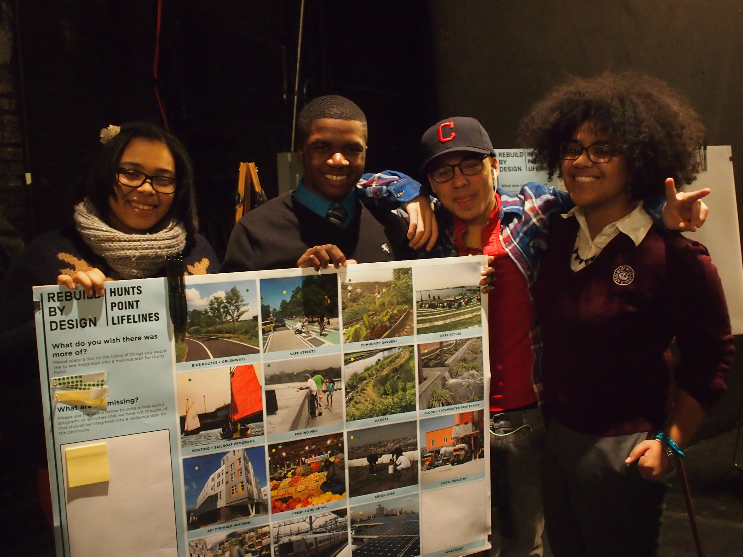 Photo of four teenagers holding up a presentation poster headed "Hunts Point Lifelines" and featuring images and headings for bike routes, greenways, safe streets, community gardens, river access and more