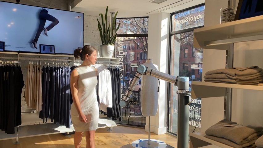 Zdjęcie kobiety ubranej w obcisłą, białą, dzianinową sukienkę wchodzącą do sklepu odzieżowego, gdzie ramię robota znajduje się w pobliżu manekina w witrynie sklepu
